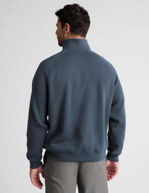 A man viewed from the back wearing a slate blue quarter-zip sweatshirt and gray trousers, set against a plain light gray studio background.