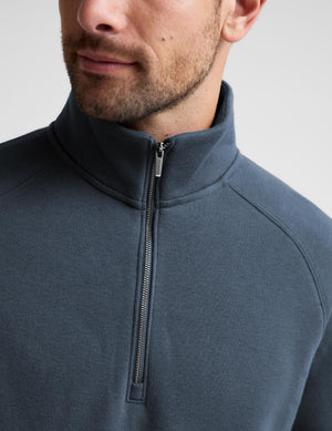 Close-up of a man wearing a dark slate blue quarter-zip pullover sweater, showing the collar, zipper, and shoulder details against a light gray background.