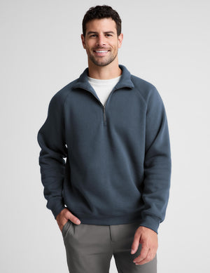 A man wearing a slate blue quarter-zip sweatshirt over a white t-shirt and gray pants, standing against a light gray studio background.