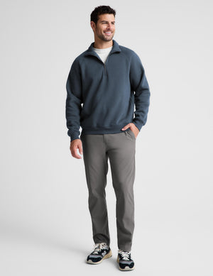 A man modeling a slate blue quarter-zip sweatshirt, gray dress pants, and navy sneakers against a light gray studio background.