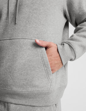 Close-up of a person wearing a light heather gray pullover hoodie, showing the front kangaroo pocket with a hand inserted, against a plain light gray background.