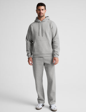 A man modeling a light gray heathered two-piece tracksuit, consisting of a pullover hoodie with a front kangaroo pocket and matching sweatpants, paired with white sneakers, against a light gray studio background.