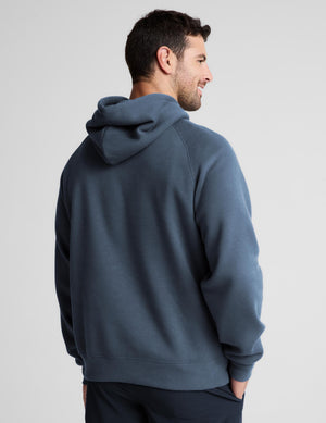 A man wearing a slate blue hooded sweatshirt, shown from a rear three-quarter view against a light gray studio background.