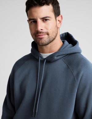 A man wearing a slate blue pullover hoodie with a white undershirt, photographed against a neutral light gray background.