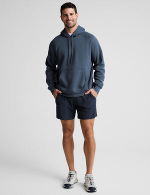 A man modeling a slate blue pullover hoodie and matching navy athletic shorts against a light gray studio background.