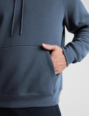 Close-up of a person wearing a slate blue-gray pullover hoodie with a front kangaroo pocket and drawstrings, with the hand in the pocket, against a light gray background.
