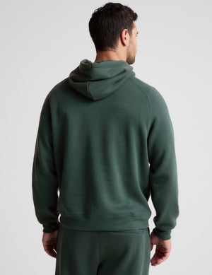 A man viewed from the back wearing a dark forest green pullover hoodie and matching sweatpants against a light gray studio background.