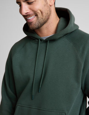 A man wearing a dark forest green pullover hoodie with matching drawstrings, shown in a close-up profile view against a neutral gray background.