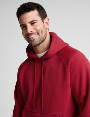 A smiling man wearing a red pullover hoodie with drawstrings, photographed against a plain light gray studio background.