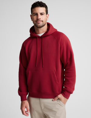 A man wearing a solid red pullover hoodie with drawstrings and a kangaroo pocket, paired with beige pants, photographed against a neutral light gray background.