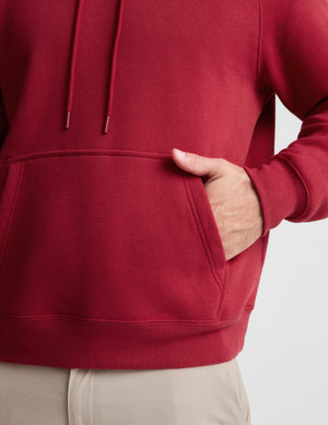 Close-up of a person wearing a red pullover hoodie with a front kangaroo pocket and drawstrings, with one hand inserted into the pocket. The person is also wearing light beige pants against a plain light gray background.