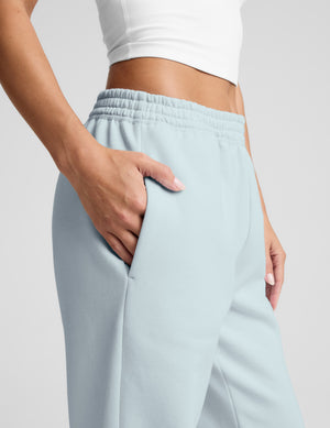 Woman wearing a white tank top and light blue sweatpants on a plain background. 