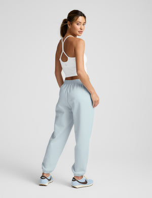 Woman wearing a white tank top and light blue sweatpants on a plain background. 
