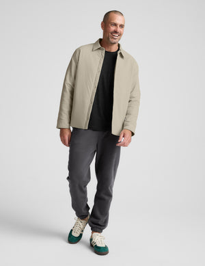 Man wearing a beige jacket, black shirt, gray pants, and white sneakers on a light gray background. 