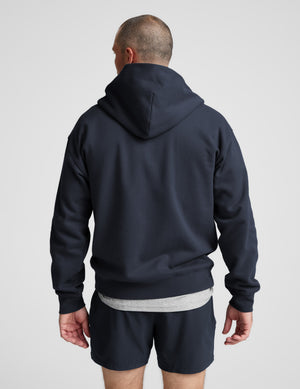 Man wearing a navy blue full zip hoodie and shorts on a plain background. 