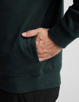 Person wearing a dark green sweatshirt with one hand in the pocket on a light gray background. 