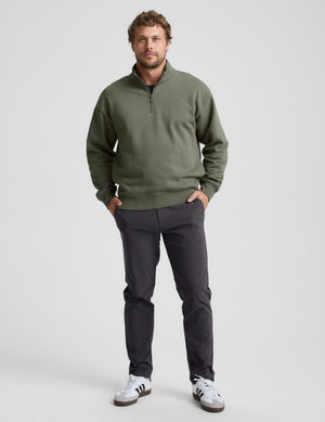 Man wearing a green quarter zip pullover and khaki pants with a plain background. 