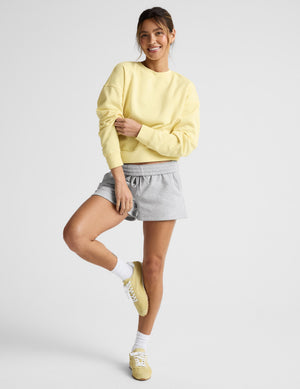 Woman wearing a yellow crew neck sweatshirt and gray shorts sitting on a plain background. 