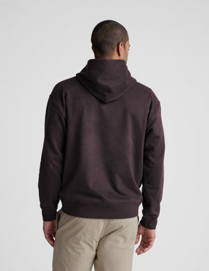Man wearing a dark brown hoodie and beige pants on a gray background. 