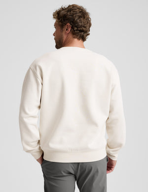 Man wearing a beige sweater and gray pants on a plain background. 