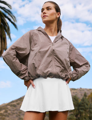 Woman wearing a brown jacket and white skirt outdoors with a blue sky and palm trees in the background. 