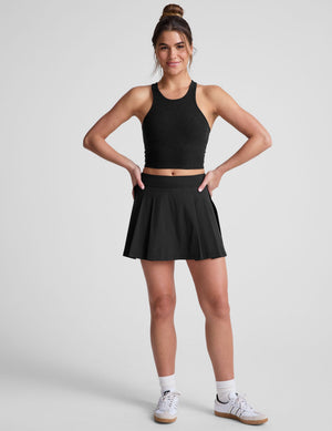 Woman wearing the black Score Pleated Skirt, a mini A-line skort with front pleats and side vents, posing with hands on hips.
