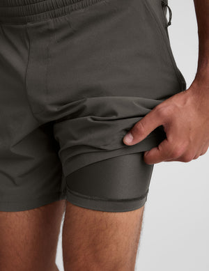 man wearing a gray short sleeve shirt and dark gray lined shorts on a plain background. 