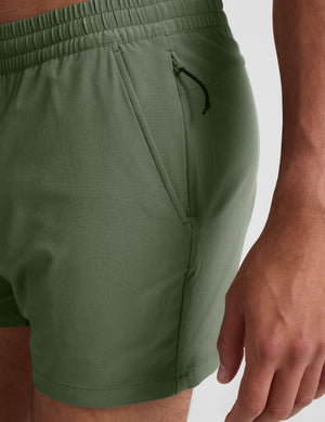 man wearing a gray t-shirt and green shorts on a plain background. 