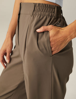 Close-up of brown Status Wide Leg Trousers with a hand in the side pocket, showing the elastic waistband and pleated front.