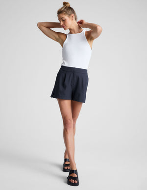 Tailored navy Status Shorts with front pleats and a mid-rise fit, shown on a model with a white tank top and sandals.