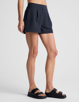 Pleated navy Status Shorts with a mid-rise waist and side slits, shown from a side profile on a model wearing sandals.