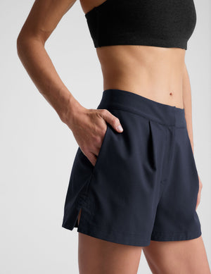 Person wearing navy Status Shorts with front pleats and a hand in the pocket. Tailored mid-rise style with a side slit.