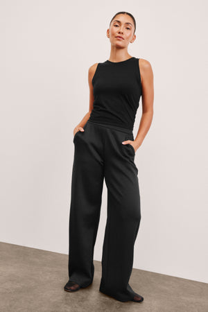 woman wearing a black sleeveless top and wide-leg pants against a plain background. 