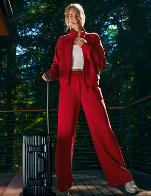 Smiling woman wears red In The Flow Piped Wide Leg Pant and matching jacket, holding a suitcase on a sunlit balcony.