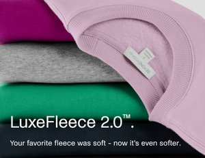 Three folded fleece sweaters in magenta, heather gray, and green are stacked diagonally, with a lavender fleece garment draped over the top right. A small fabric tag is visible on the collar.