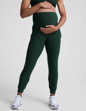 model wearing green maternity midi length joggers with pockets on a plain background. 