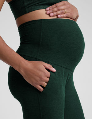 model wearing green maternity midi length joggers with pockets on a plain background. 