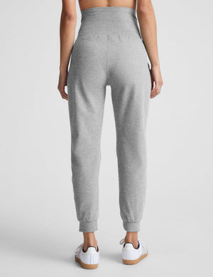 Pregnant woman wearing a white tank and gray joggers against a plain background. 