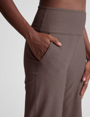 Woman wearing a white bra and brown wide leg pants on a light gray background. 