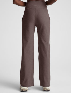 Woman wearing a white bra and brown wide leg pants on a light gray background. 