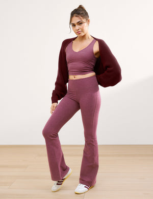 Woman wearing a burgundy shrug and purple v-neck cropped tank and mid-rise bootcut pants against a plain background. 