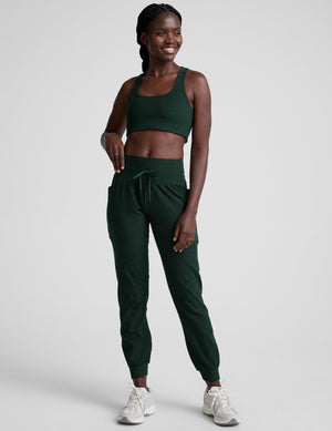 model wearing a green sports bra and green midi length joggers with a drawcord at the waistband against a plain background. 
