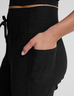 model wearing black high-waisted midi length joggers with a drawcord at waistband and pockets against a plain background. 