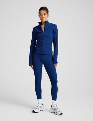 Woman wearing a blue zip-up athletic jacket and leggings on a light background. 