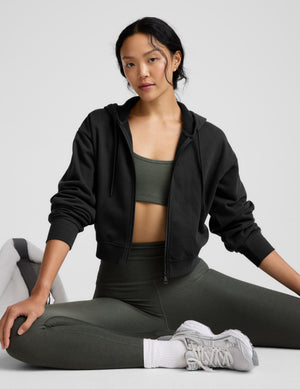 Woman in black hoodie and green leggings sitting on a white surface with a neutral background. 