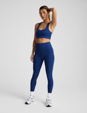 Woman wearing a blue sports bra and high-waisted midi length leggings against a plain background. 