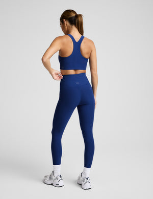 Woman wearing a blue sports bra and high-waisted midi length leggings against a plain background. 