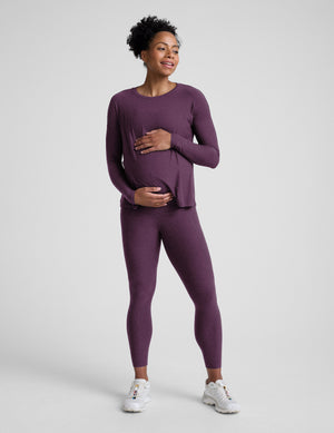 Woman wearing a purple maternity long sleeve and pocket leggings against a plain background. 