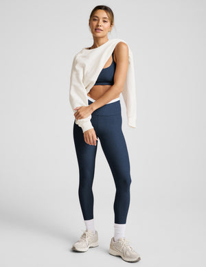 Woman wearing navy leggings, sports bra, and white sneakers on a light gray background. 