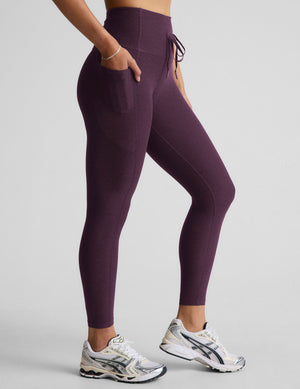 Woman wearing purple midi length leggings with a drawcord at waistband on a gray background. 